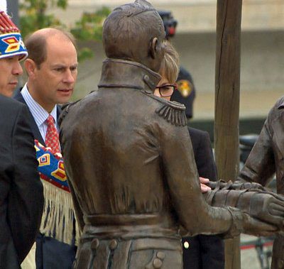 Indigenous man with head dress with white man looking at metal statues of European and Indigenous men exchanging blanket between their hands
