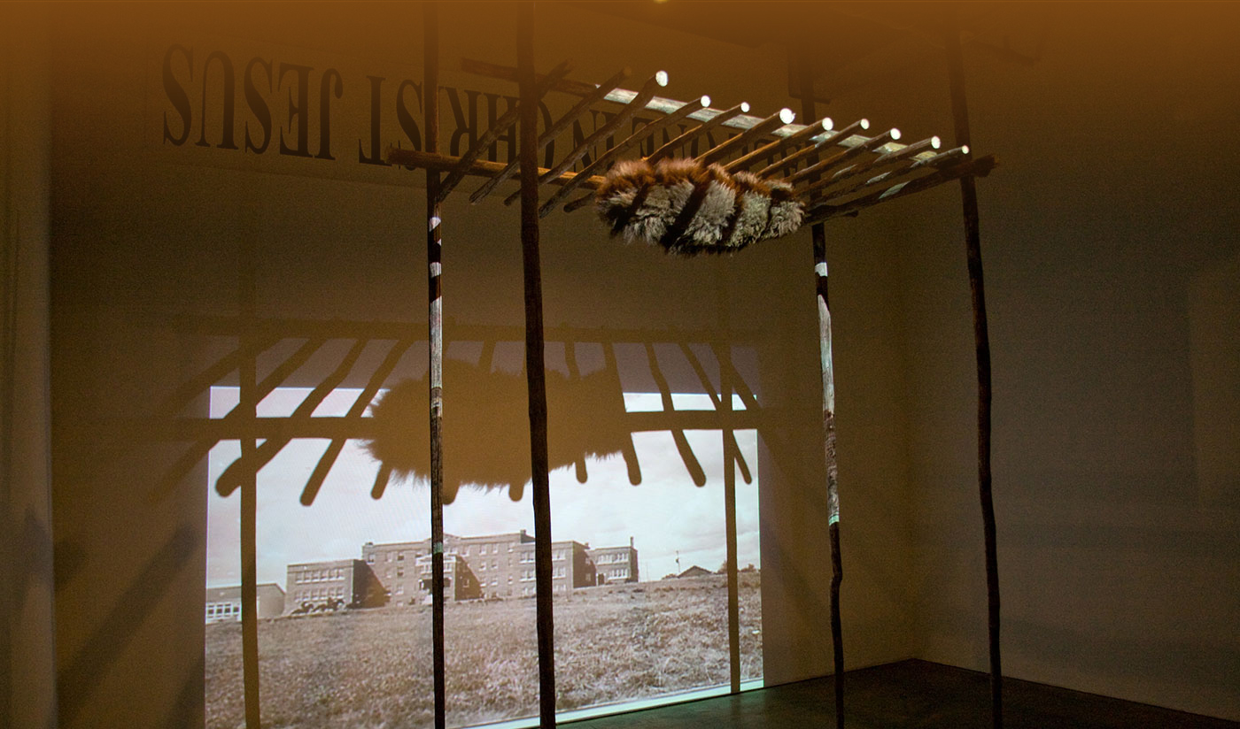 Structure made of wooden stakes holding up a bison hide in shape of person in a blanket in front of wall with projection of archival image of residential school