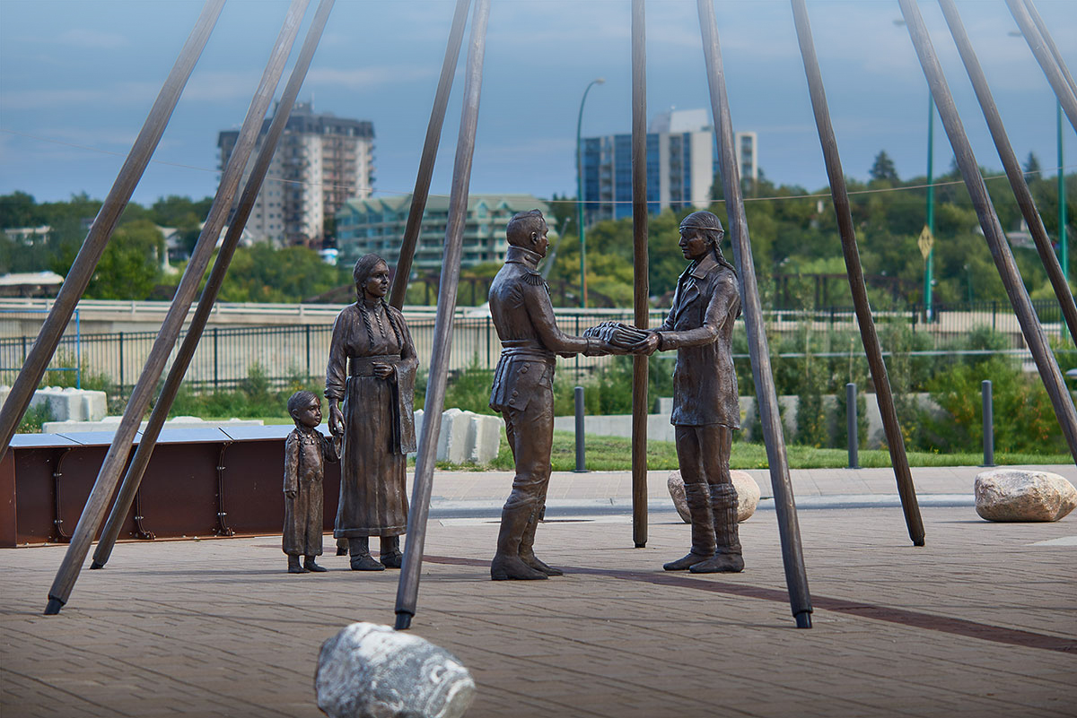 Four metal statues form a group under the metal framework of a teepee's frame, residential apartment towers and trees indicate an urban setting. The statues, from left to right, a small Indigenous child in traditional dress and braids holds the hand of a woman dressed similarly also with braids; European man in colonial dress exchanging blanket handed to him from Indigenous man in mix of colonial and traditional native dress, his hair in braids.