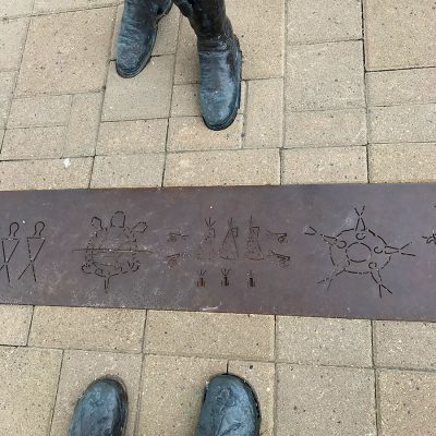 Metal strip on ground with etched in figures. The strip is between interlocking stones. On either side are the feet of metal statues, top in colonial boots, bottom in traditional native boots with beadwork tracings.